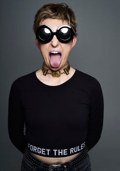 Studio portrait of a young woman with blonde hair wearing large black sunglasses and mauve lipstick sticking her tongue out at the camera