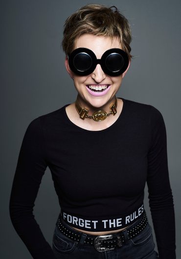 Studio portrait of a young woman with blonde hair wearing large black sunglasses and mauve lipstick laughing at the camera