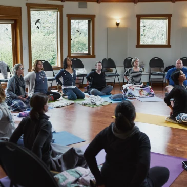 A yoga instructor sits in the middle of a room demonstrating a heart opening pose while leading a yoga session at a self care for educators workshop at Roundhouse Farm, Victoria BC